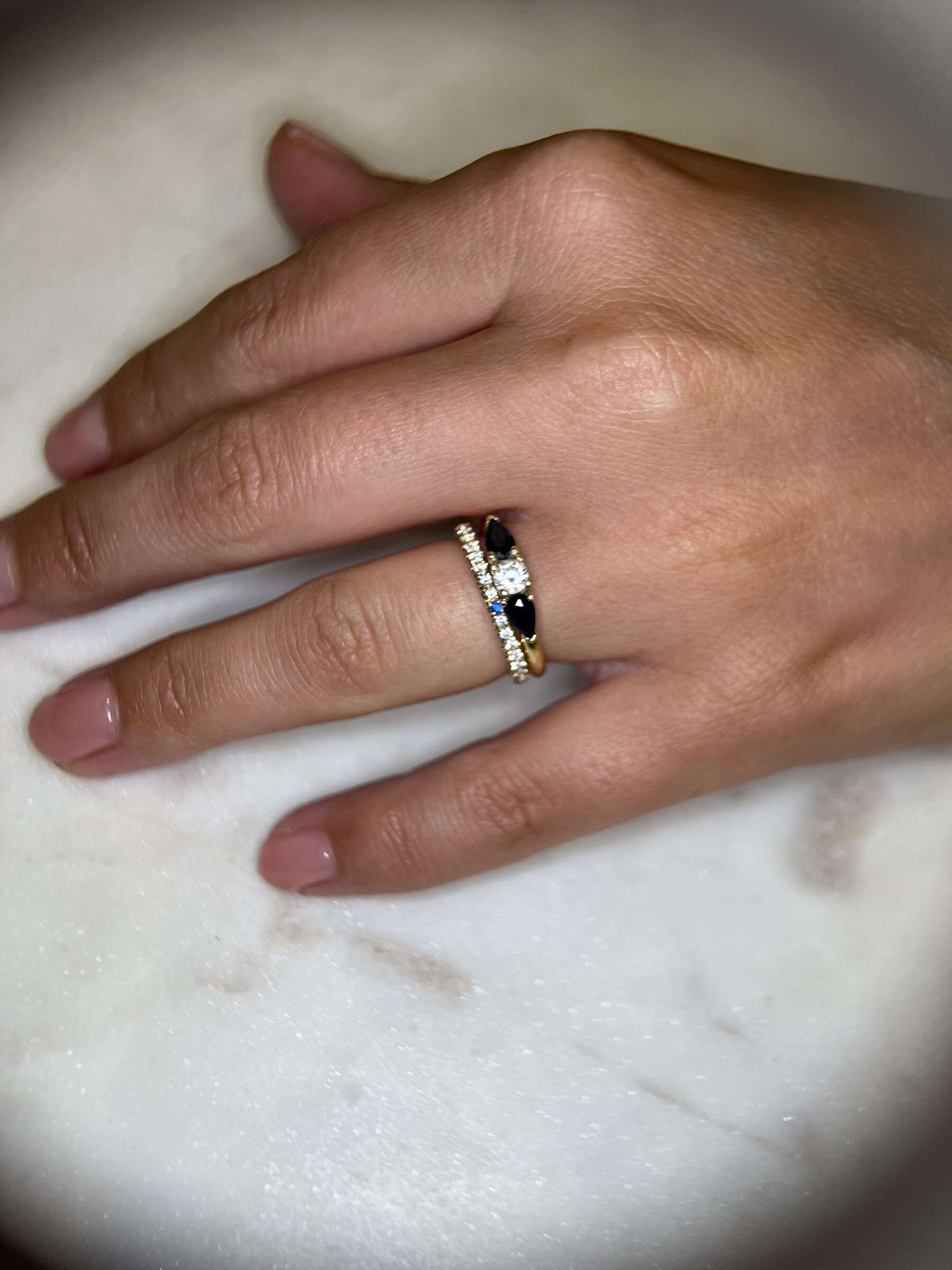 Hand met verlovingsring met diamant en twee druppelvormige saffieren, naast een diamanten ring